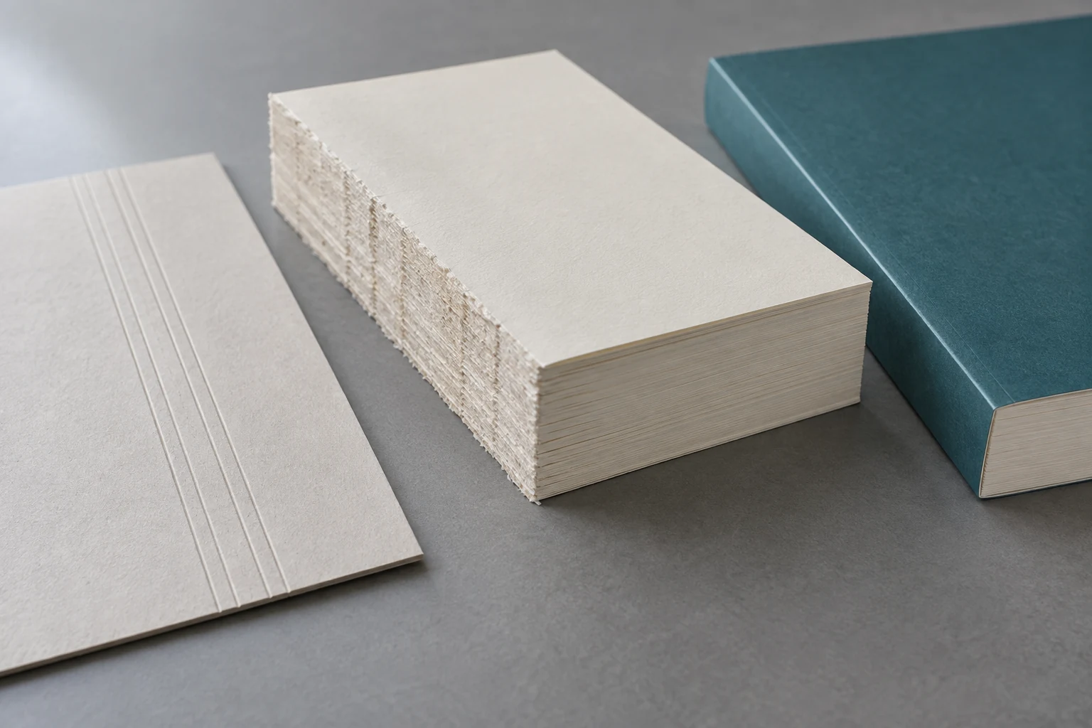 Roughened cream book-block spine beside a scored cover sheet and a teal perfect-bound paperback