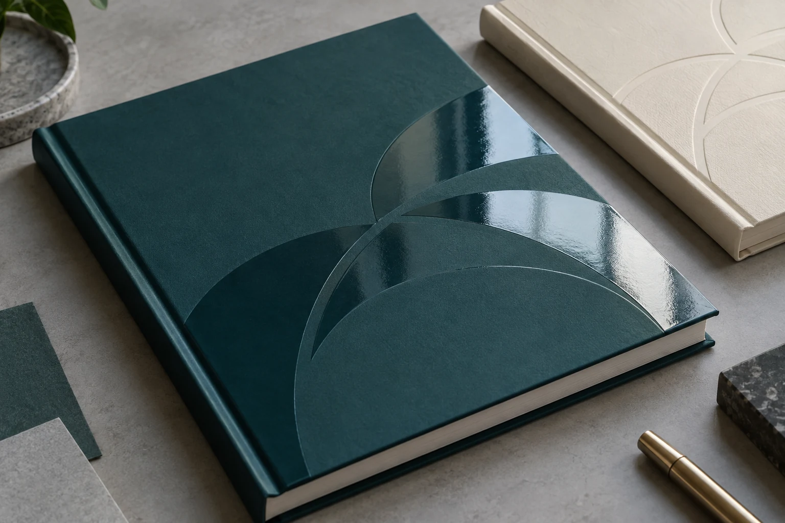 Glossy clear spot UV arcs on a matte teal hardcover beside an ivory blind relief cover sample